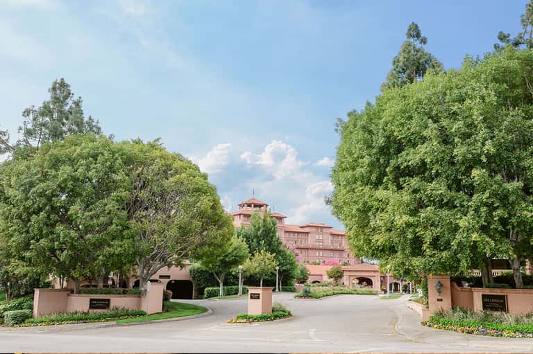 Tree-lined driveway entrance to pink Mediterranean-style mansion with manicured grounds