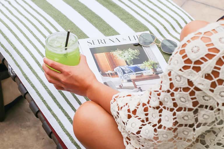 Woman relaxing poolside with green drink, magazine, sunglasses, and striped lounge chair