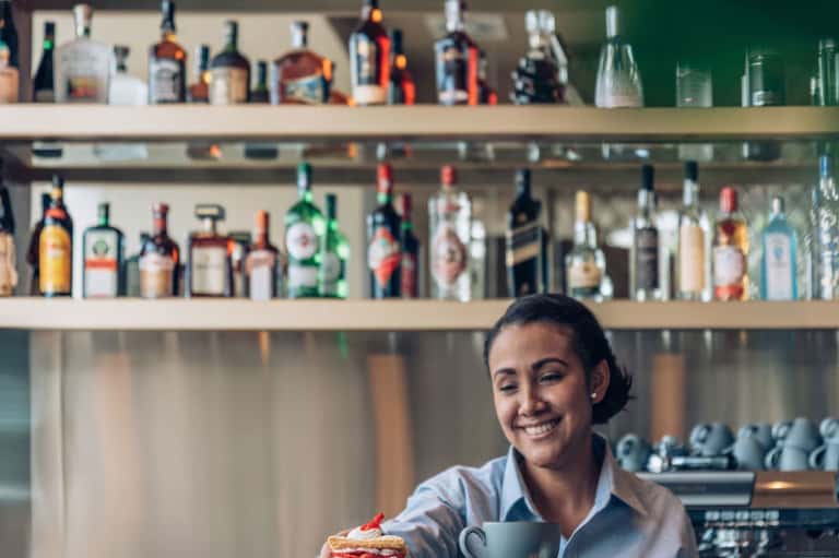 Smiling bartender in white shirt serving dessert at hotel bar with shelves of liquor bottles behind
