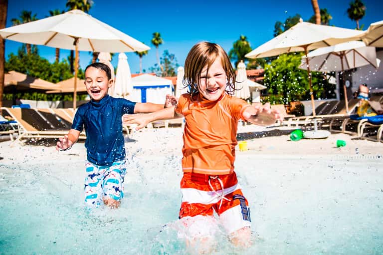 Two children playing and splashing in a shallow resort pool with umbrellas and palm trees in background
