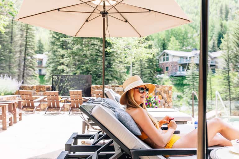 Woman relaxing on lounge chair under beige umbrella at resort pool with forest backdrop