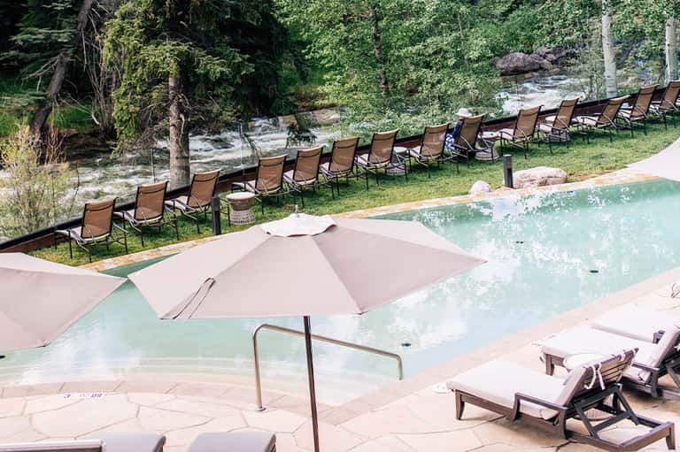 Natural pool surrounded by forest, lounge chairs, umbrellas, and creek with lush greenery