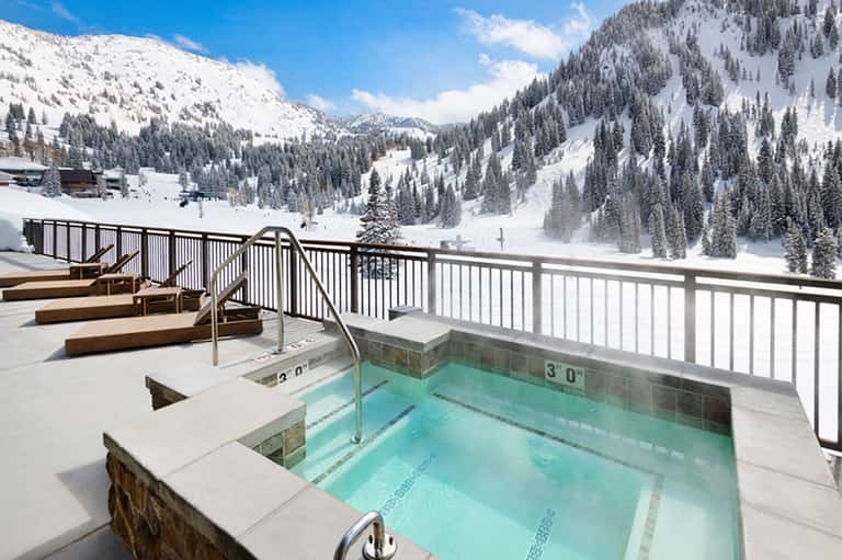 Outdoor hot tub on snowy mountain terrace with white railings and forested peaks in background