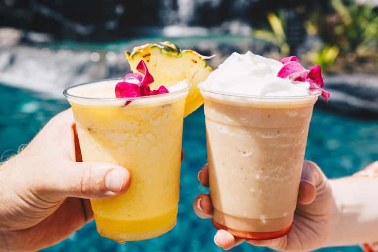 Two tropical cocktails with pineapple and flower garnishes held by poolside with water view