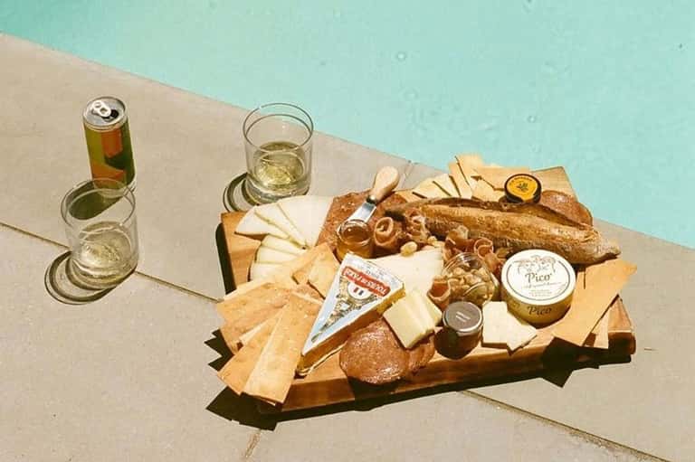Poolside charcuterie board with cheeses, bread, and beverages beside turquoise water