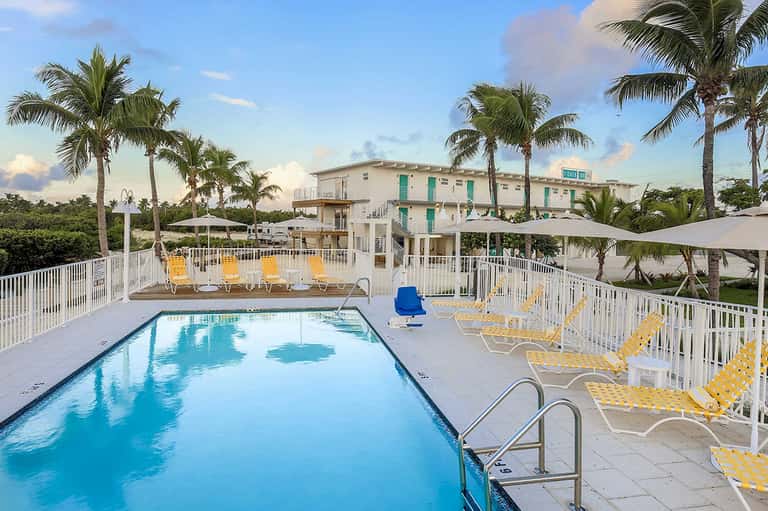 Resort pool with yellow loungers, palm trees, white railing, and beach building in background