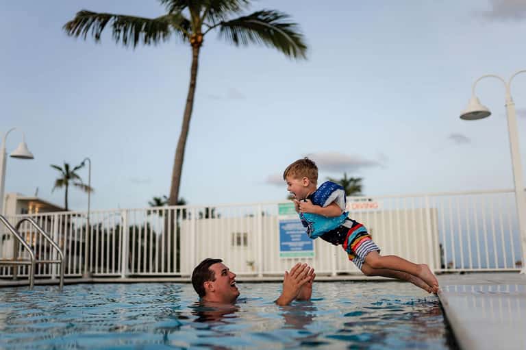 Father playing with young boy in resort pool with palm tree and white railing backdrop