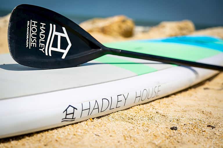 Paddle board with Hadley House logo on sandy beach near ocean rocks