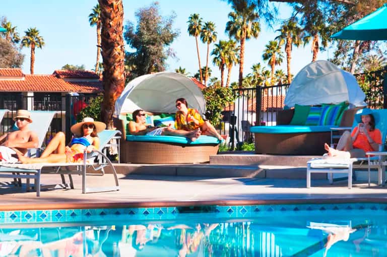 Resort pool area with lounge chairs, cabanas, and palm trees under clear blue sky