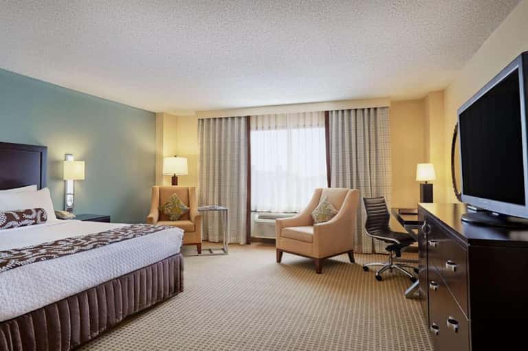 Hotel bedroom with queen bed, beige armchair, flat-screen TV, and window with drapes