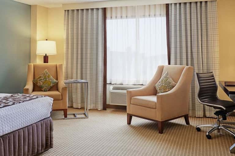 Hotel suite with queen bed, seating area, desk, and view of checkered curtains and window