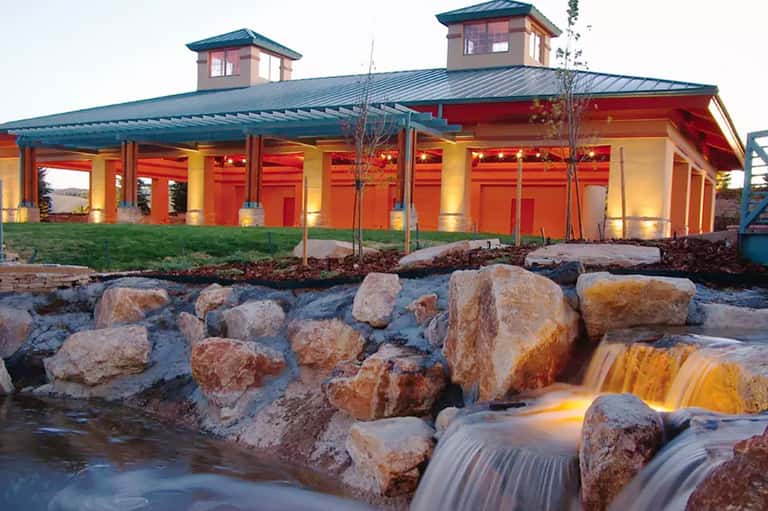 Red and yellow modern building with two cupolas beside illuminated waterfall and rocky stream at dusk