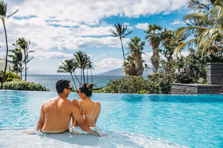 Couple relaxing in infinity pool with ocean view, palm trees, and mountains in background
