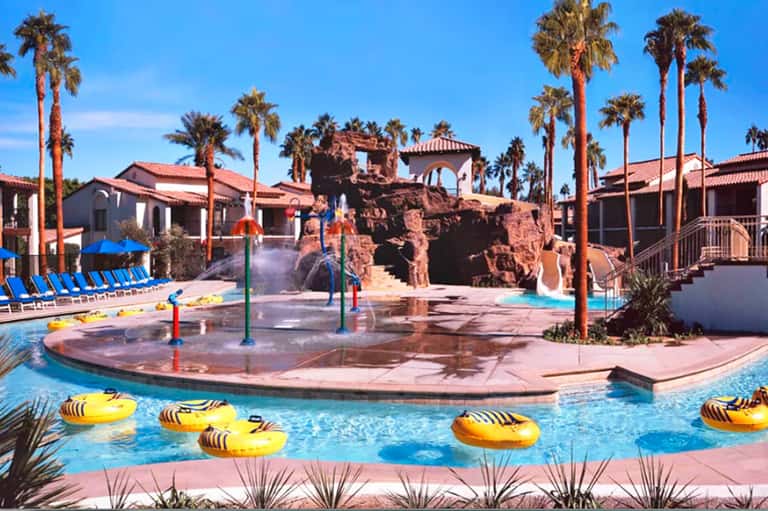 Resort water park with artificial rock formations, slide, and yellow inflatable floats surrounded by palm trees