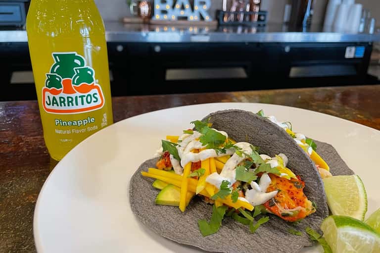 Two charcoal fish tacos with yellow mango strips, lime wedge, and Jarritos pineapple soda on bar counter