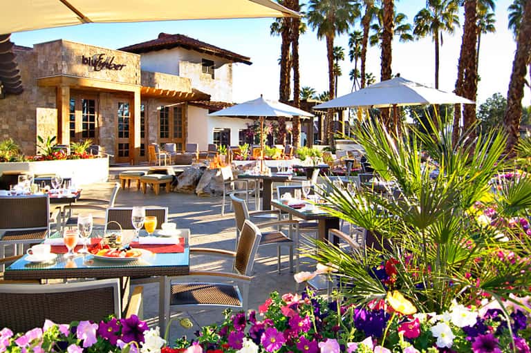 Outdoor resort dining area with colorful flowers, umbrellas, and palm trees in sunny setting
