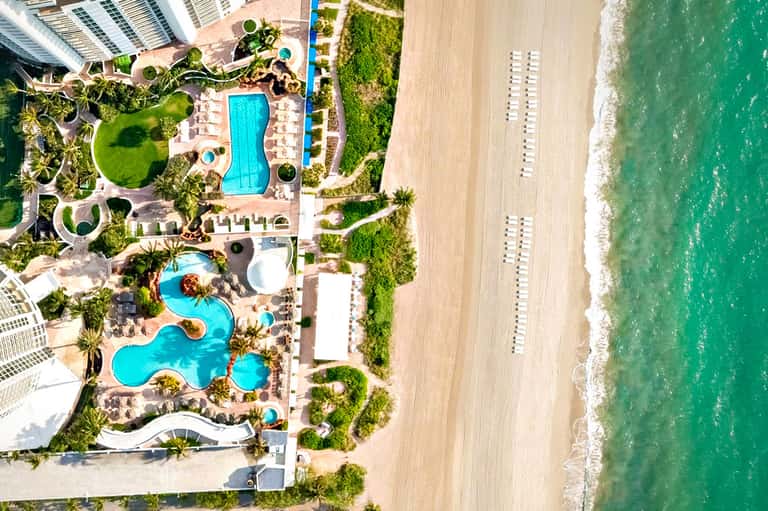 Overhead view of resort pool complex with lounge chairs, beach access, and green landscaping along shore