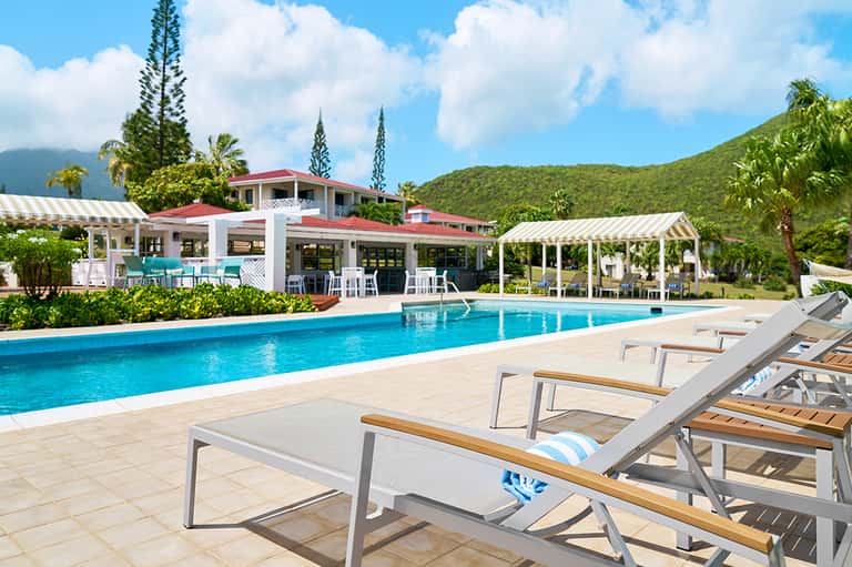 Resort pool with mountain backdrop, lounge chairs, cabanas with red roof, and tropical landscaping
