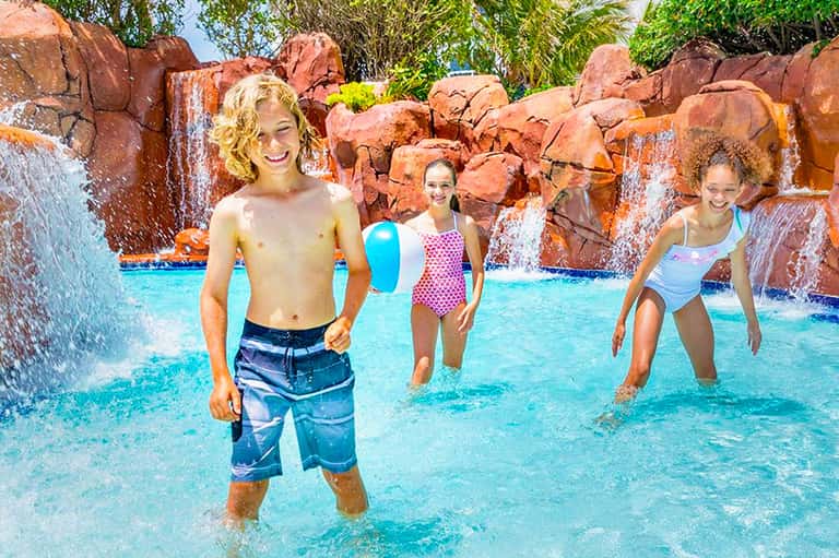 Children playing in a bright turquoise wave pool with red rock formations and waterfalls