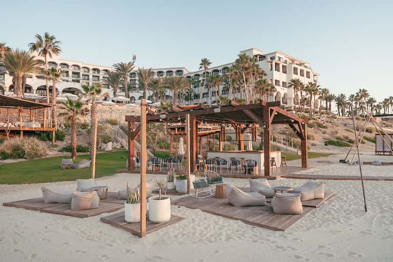 Beachfront resort with palm trees, wooden pergola structures, lounge seating, and hotel buildings