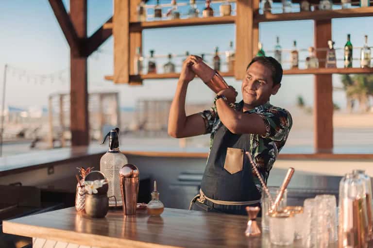 Bartender preparing tropical cocktail behind beachfront bar with ocean view in background