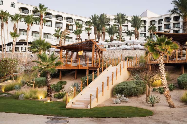 Beachfront resort with white buildings, wooden boardwalk, desert landscaping, and palm trees