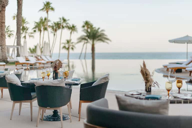 Beachfront dining area with palm trees, ocean view, elegant table settings, and white lounge furniture