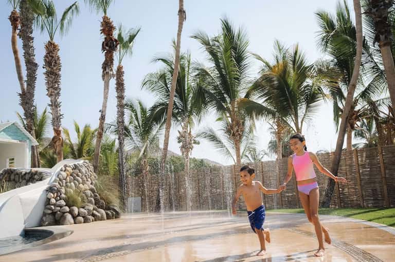 Children playing in shallow splash pad surrounded by palm trees and resort buildings