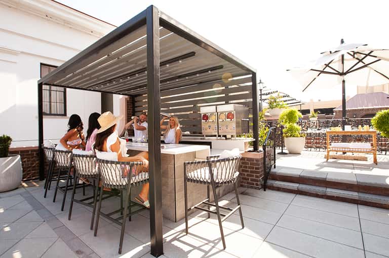 Rooftop bar with pergola structure, patrons at counter, city skyline views, and umbrella seating
