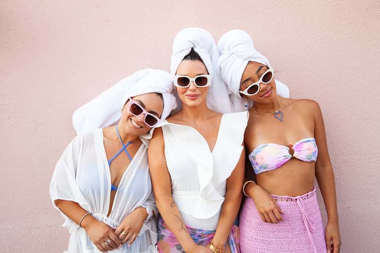Three women in beach attire with white sunglasses and wrapped towels