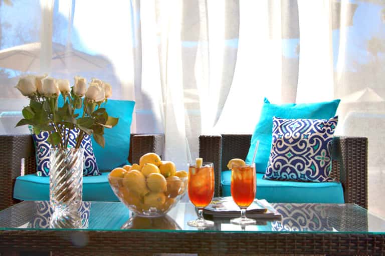 Turquoise lounge seating with patterned pillows, fresh flowers, tropical drinks, and citrus fruit on glass table