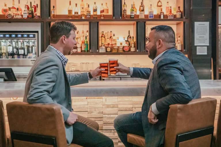 Two men toasting cocktails at upscale bar with extensive liquor collection and wooden shelving