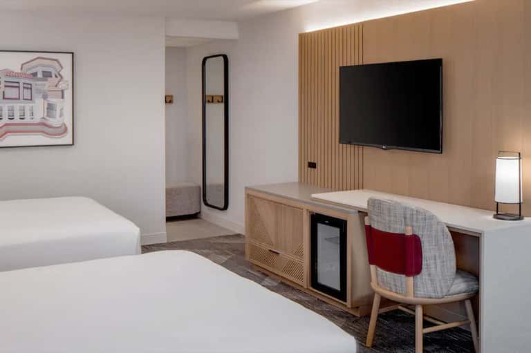 Modern hotel room with two beds, desk area, wall-mounted TV, and natural wood accents