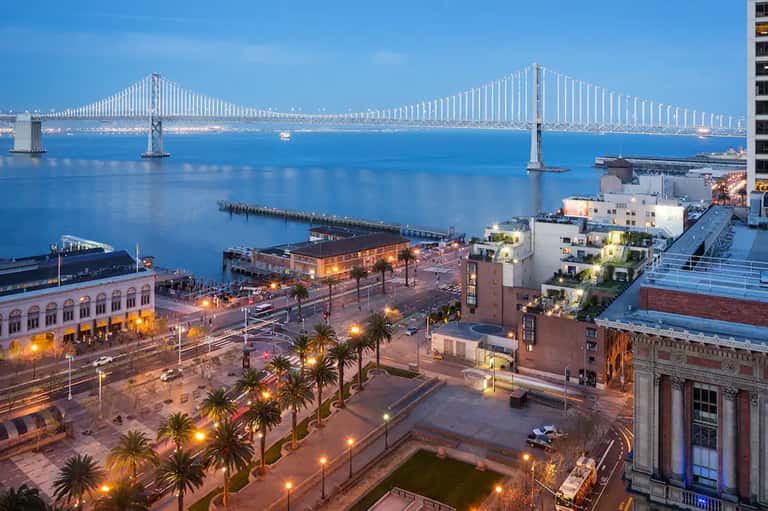 Bay Bridge and waterfront cityscape at dusk with palm-lined promenade and lights