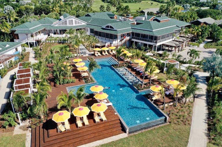 Aerial view of resort with curved pool, yellow umbrellas, palm trees, wooden decks, and surrounding tropical landscape