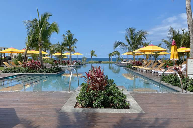 Resort pool with yellow umbrellas, loungers, palm trees, and ocean view in background