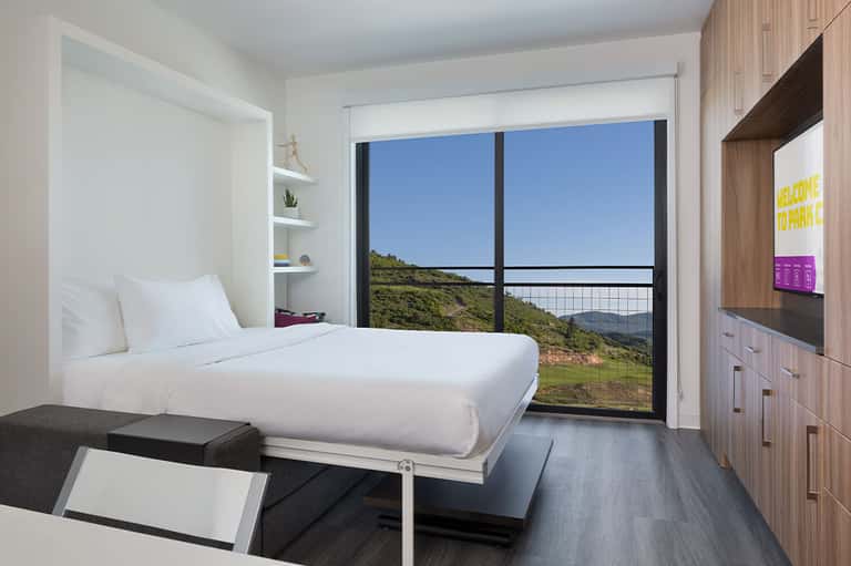 Modern hotel room with white bedding, balcony overlooking green hillside landscape
