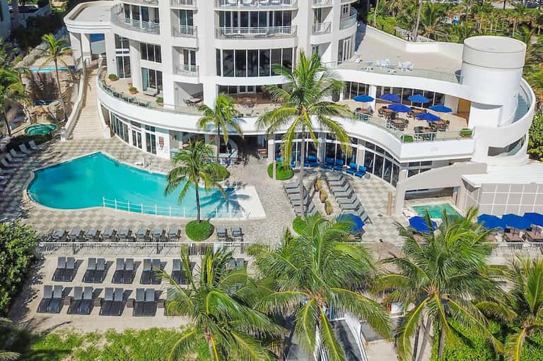 Aerial view of resort with curved white building, turquoise pool, palm trees, and blue umbrellas