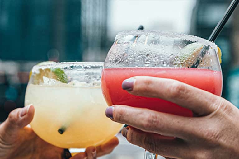 Two colorful tropical cocktails with ice and garnish held by person at bar