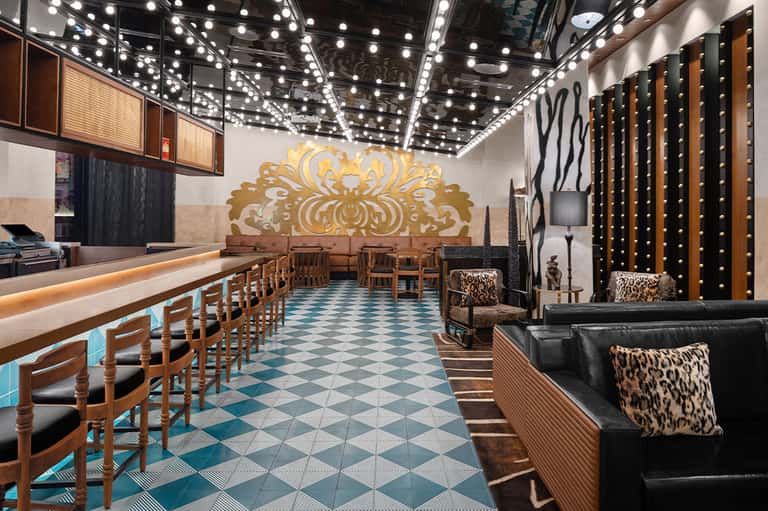 Stylish hotel bar with ornate gold wall art, teal checkered flooring, wooden chairs, and leopard print accents