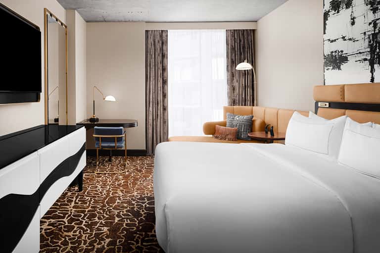 Contemporary hotel guest room with tan leather headboard, white bedding, dark TV, and patterned carpet