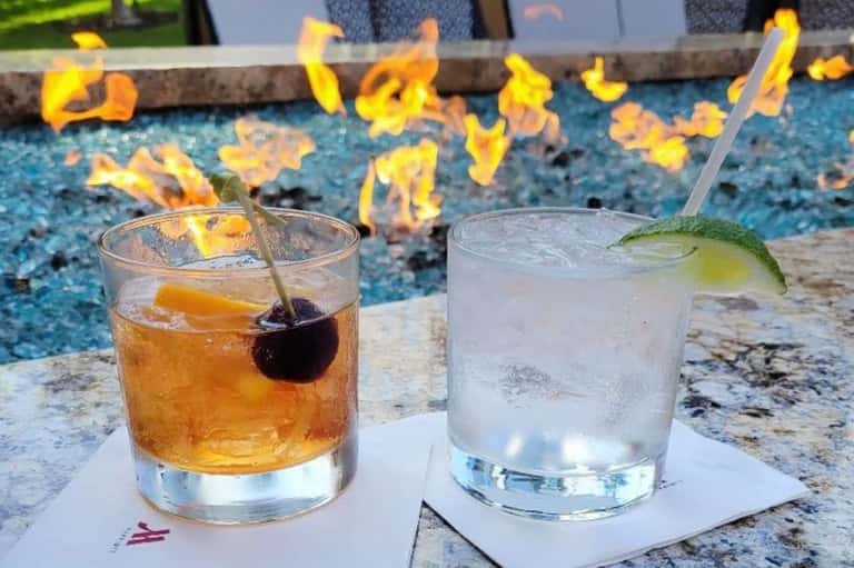 Two poolside cocktails on granite edge - amber drink and clear drink with fire pit and pool background