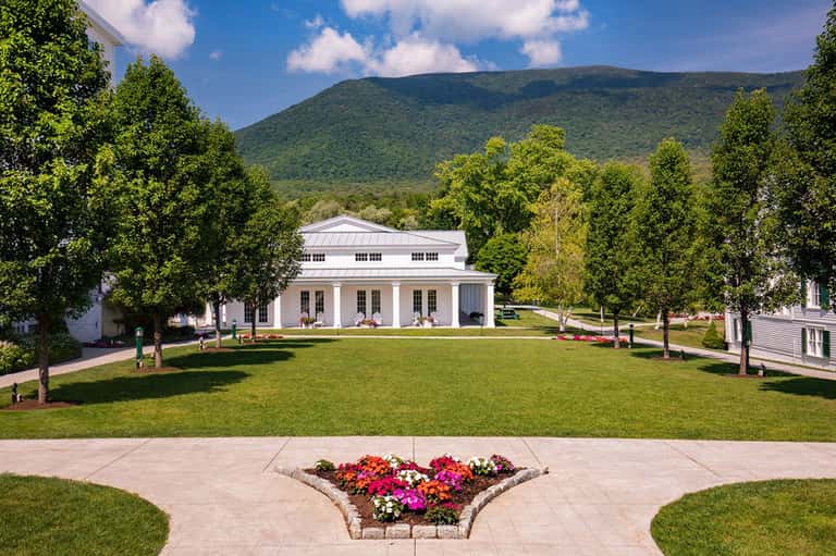 White classical building with columns surrounded by manicured lawn and mountains in background