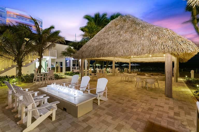 Oceanfront patio at dusk with fire pit, Adirondack chairs, thatched-roof gazebo, and palm trees