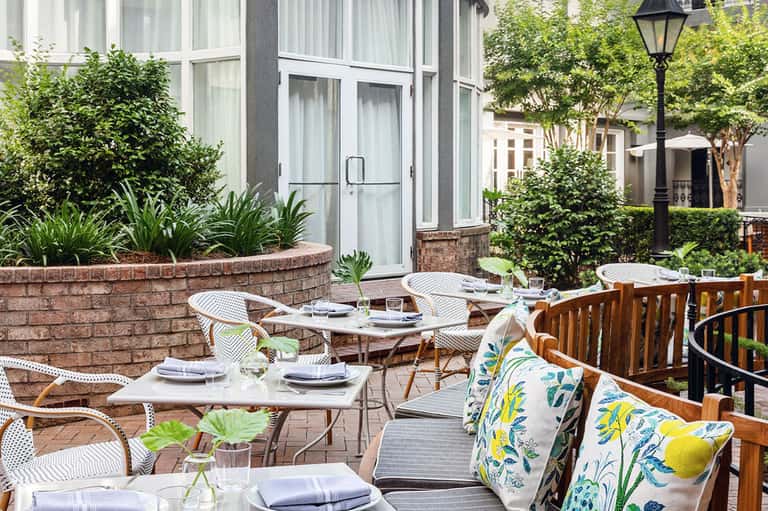 Outdoor courtyard dining space with white chairs, green landscaping, and brick planter walls