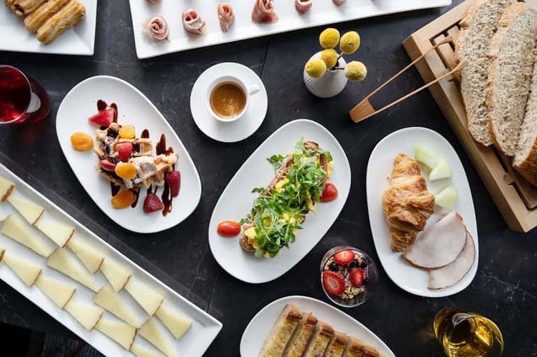 Fine dining breakfast spread with pastries, meats, fresh fruit, and espresso on dark table