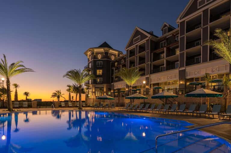 Illuminated beachfront resort at dusk with lit pool, palm trees, and waterfront buildings
