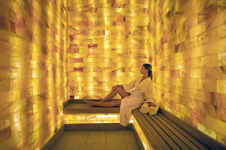 Woman in spa robe relaxing in golden salt therapy room with illuminated walls