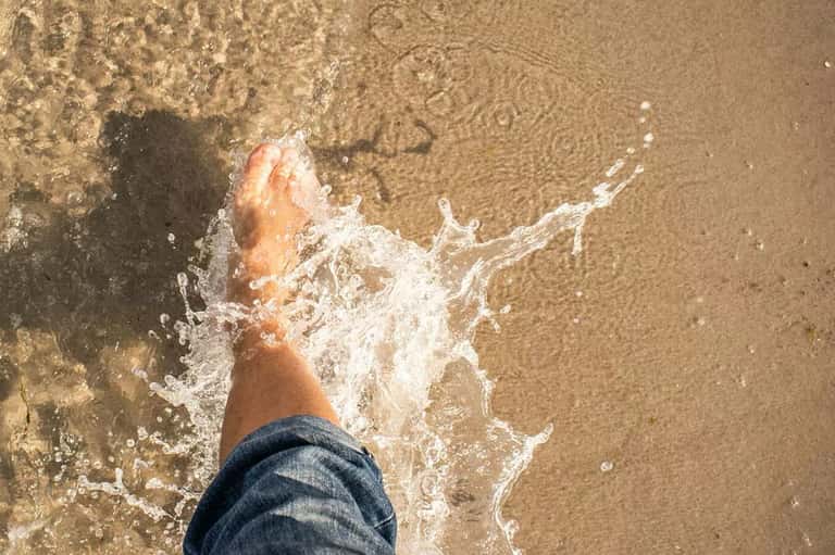 Close-up of feet in ocean waves washing onto sandy beach