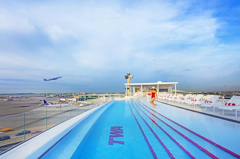 Rooftop swimming pool with lane markers overlooking airport runway, airplanes, and blue sky.